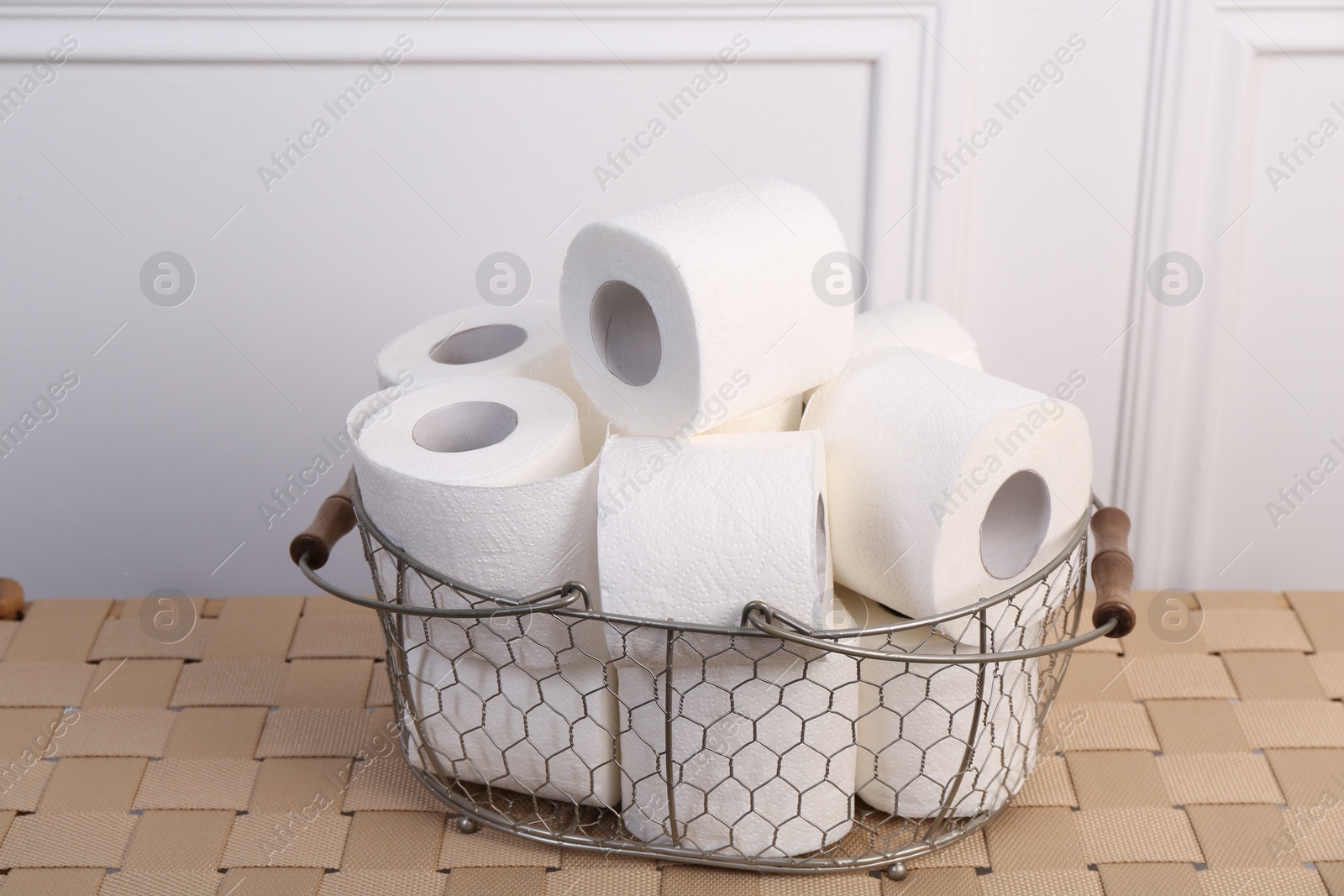 Photo of Soft toilet paper rolls in metal basket on wicker table, closeup