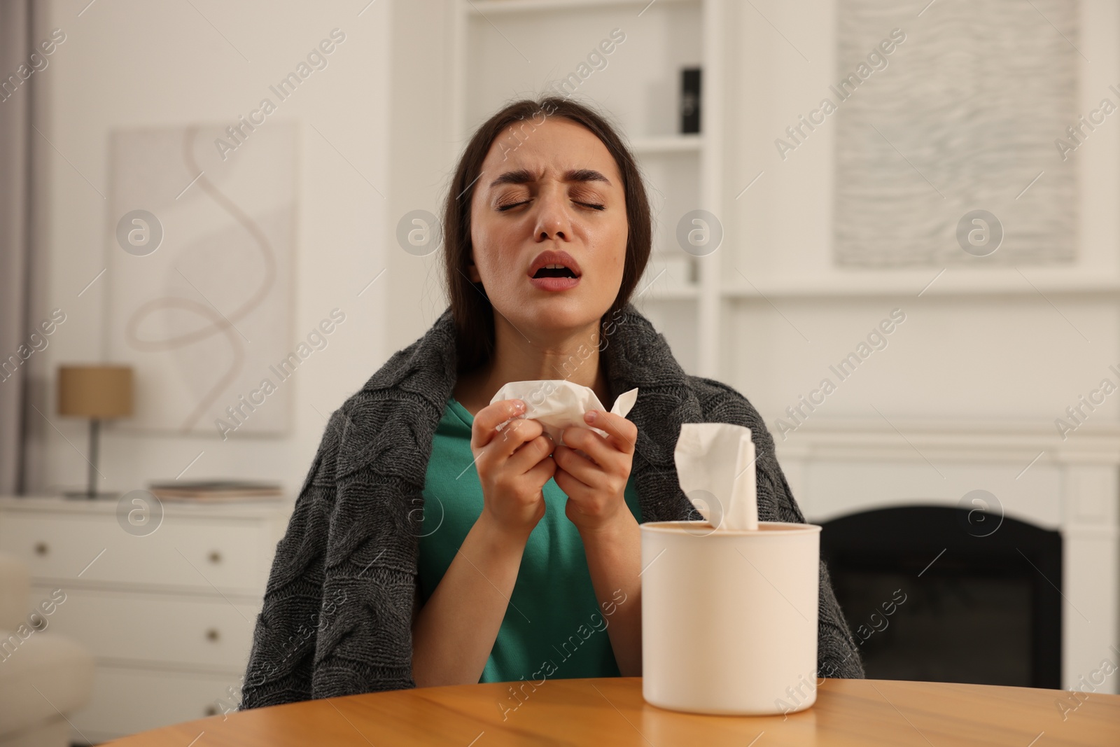 Photo of Sick woman wrapped in blanket with tissue at wooden table indoors. Cold symptoms