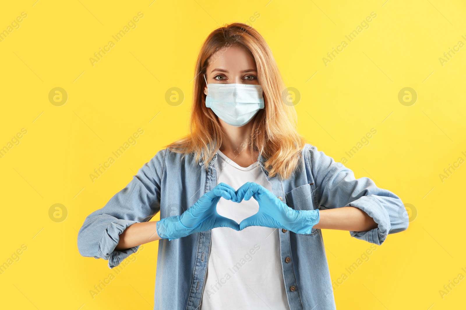 Photo of Young woman in medical gloves and protective mask making heart with hands on yellow background