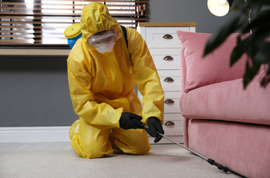 Photo of Pest control worker in protective suit spraying insecticide under sofa at home