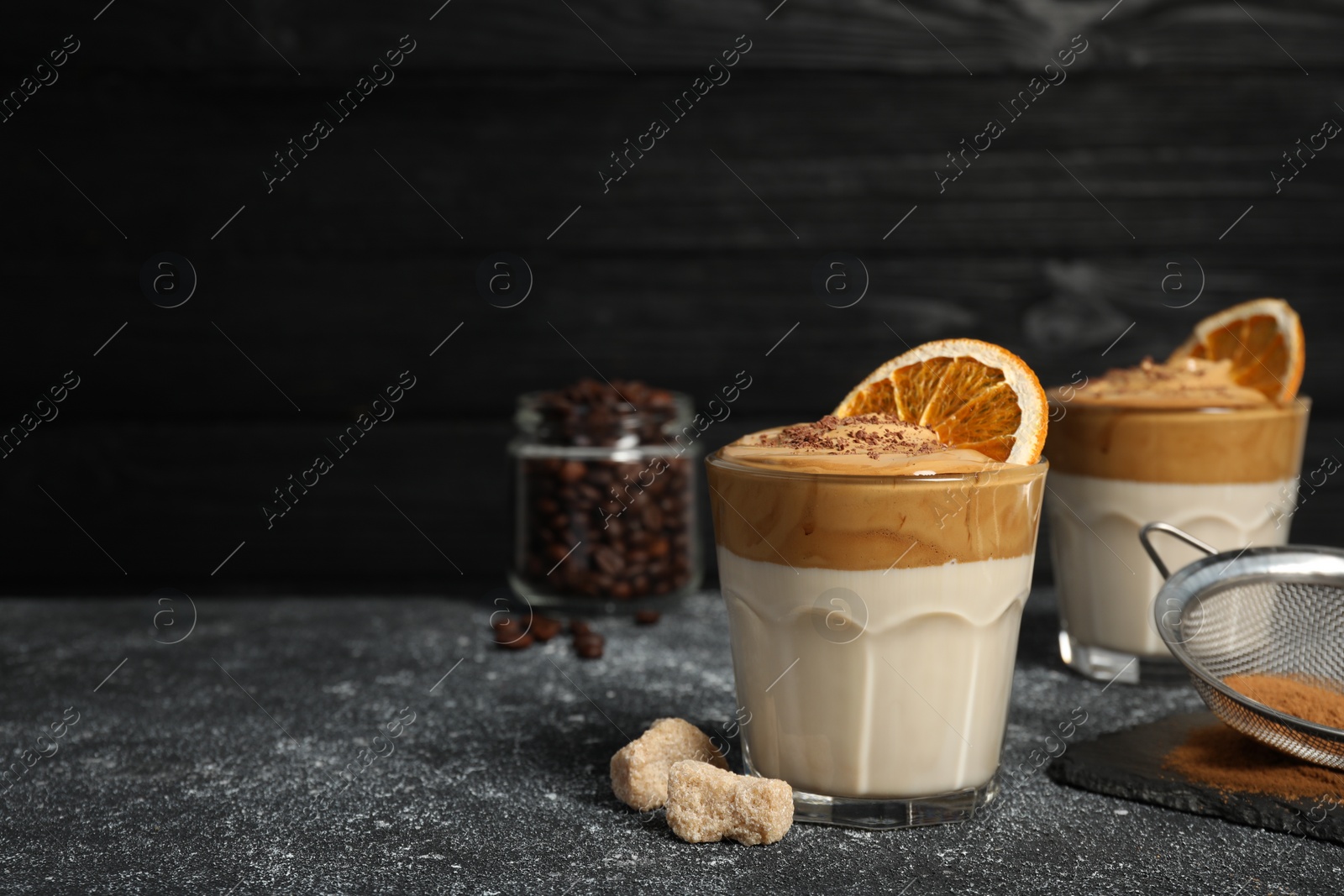 Photo of Glasses of delicious dalgona coffee with dry orange, sugar and chocolate on grey textured table, space for text