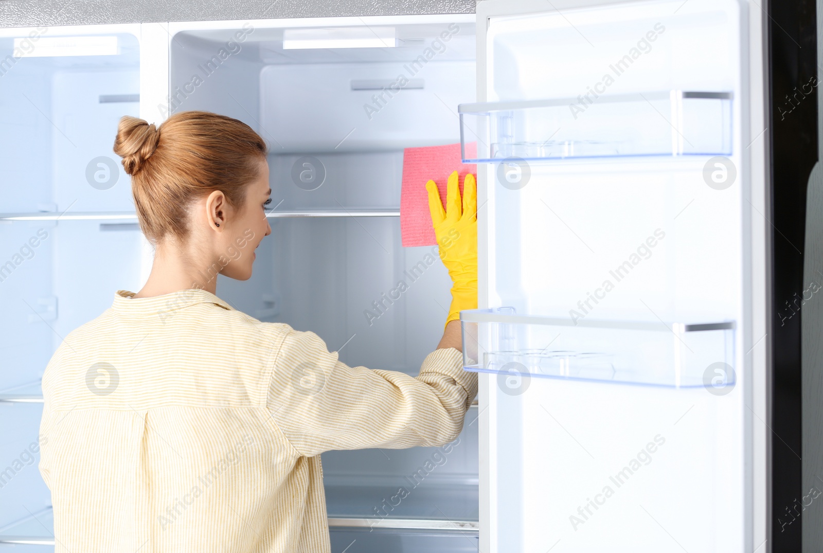 Photo of Woman in rubber gloves cleaning empty refrigerator with rag at home