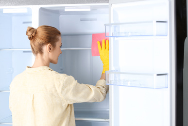 Photo of Woman in rubber gloves cleaning empty refrigerator with rag at home