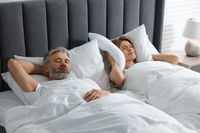 Photo of Irritated woman covering her ears with pillows in bed at home. Problem with snoring husband