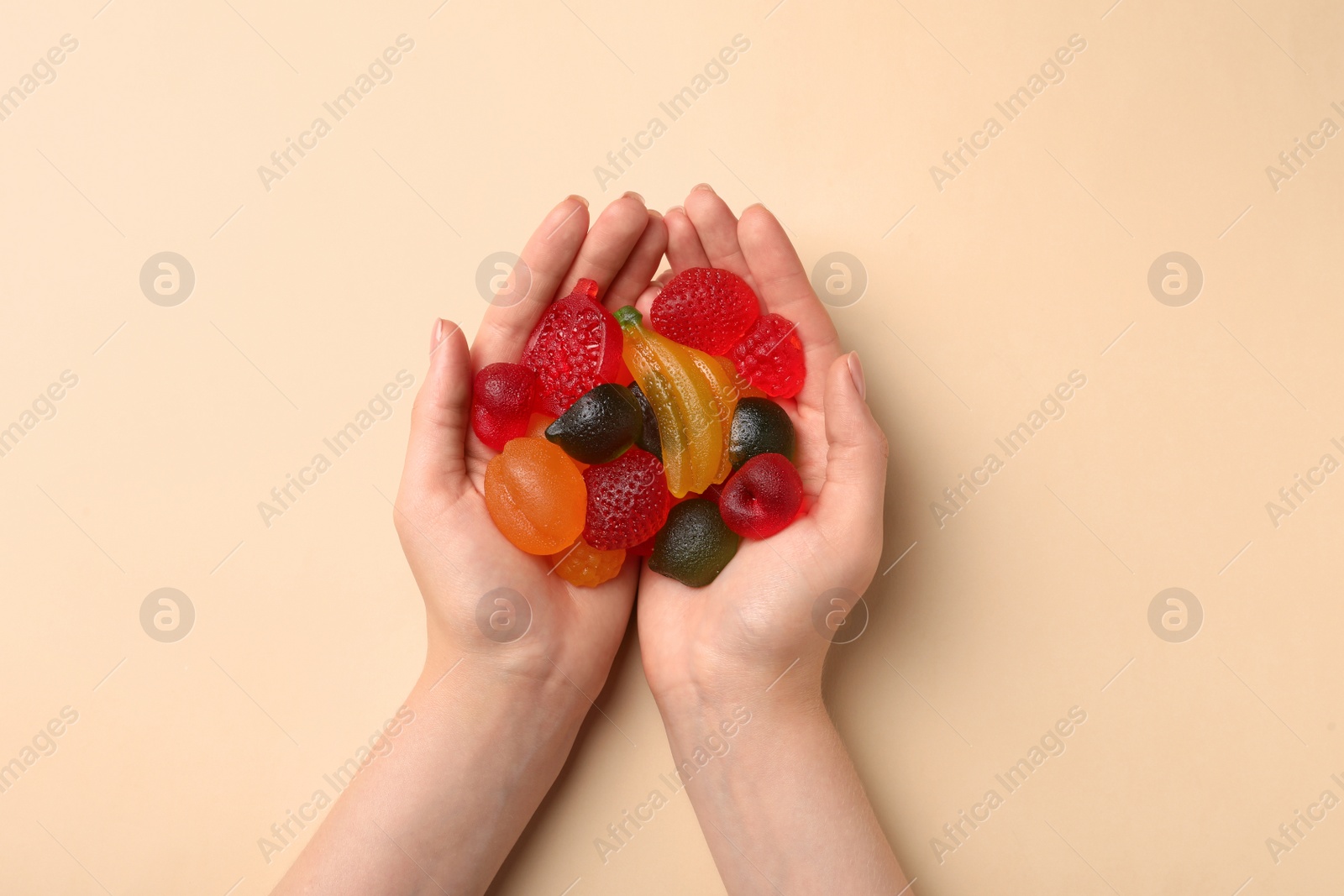 Photo of Woman holding pile of delicious fruit shaped gummy candies on beige background, top view