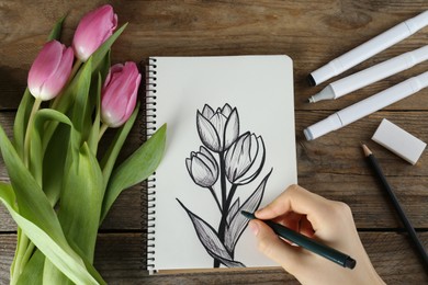 Woman drawing beautiful tulip flowers in sketchbook at wooden table, top view