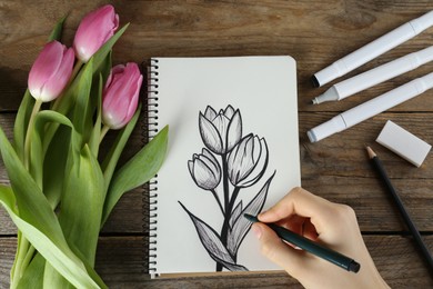 Photo of Woman drawing beautiful tulip flowers in sketchbook at wooden table, top view