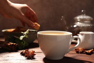 Photo of Woman taking cookie from bowl at wooden table, focus on cup of anise tea with milk