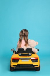 Photo of Cute little girl with toy bear driving children's car on light blue background, back view