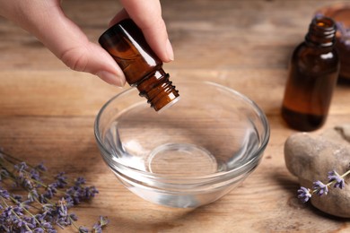 Photo of Woman dripping lavender essential oil from bottle into bowl at wooden table, closeup