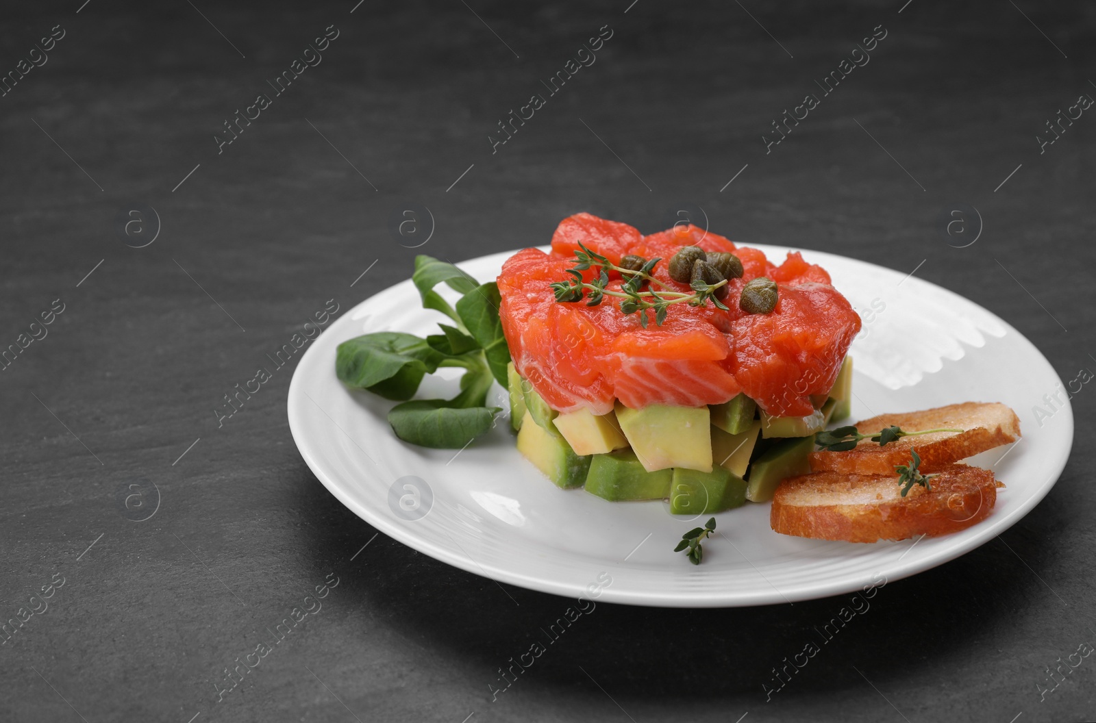 Photo of Delicious salmon tartare served with croutons, capers and greens on dark table. Space for text