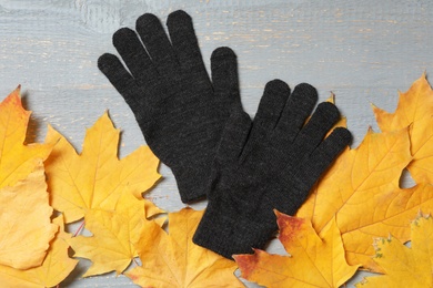 Stylish black woolen gloves and dry leaves on grey wooden table, flat lay