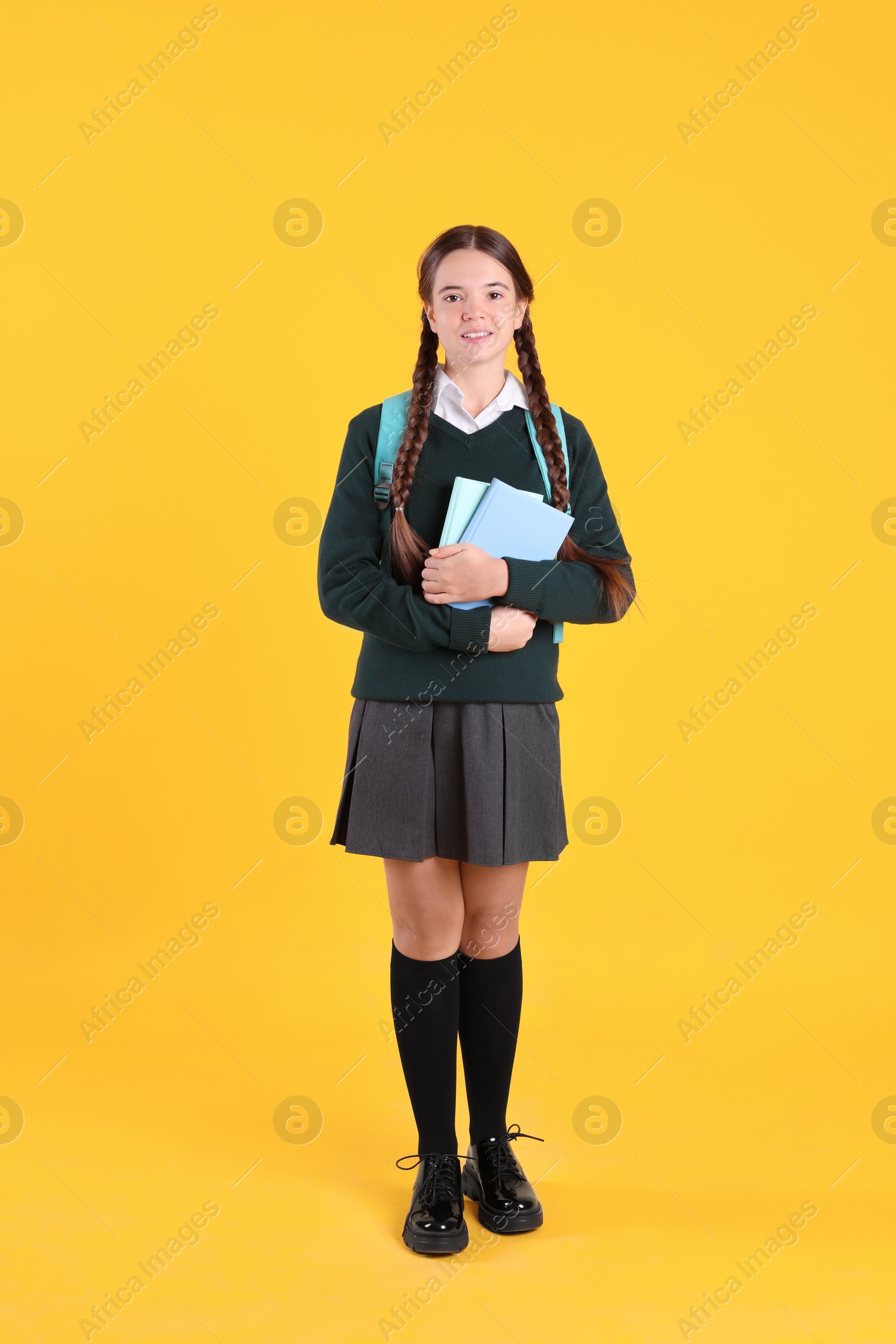 Photo of Teenage girl in school uniform with books and backpack on yellow background