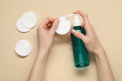 Woman applying makeup remover onto cotton pad on beige background, top view