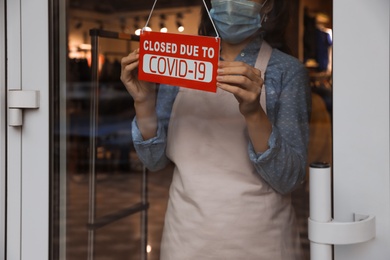 Photo of Woman in mask putting red sign with words "Closed Due To Covid-19" onto glass door, closeup