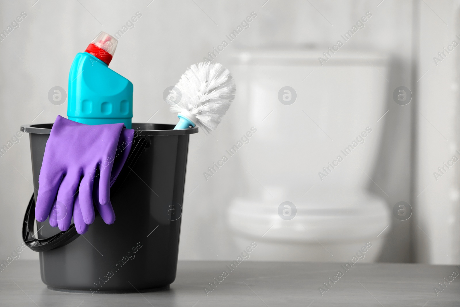 Photo of Bucket with bottle, toilet brush and gloves on table indoors, space for text. Cleaning supplies