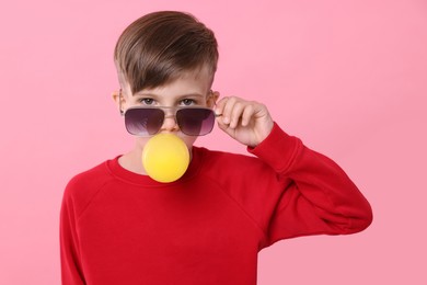 Photo of Boy in sunglasses blowing bubble gum on pink background