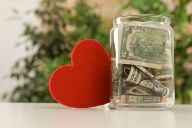 Photo of Red heart and donation jar with money on table against blurred background