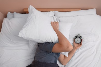 Photo of Man covering his face with pillow and holding alarm clock in bed, above view