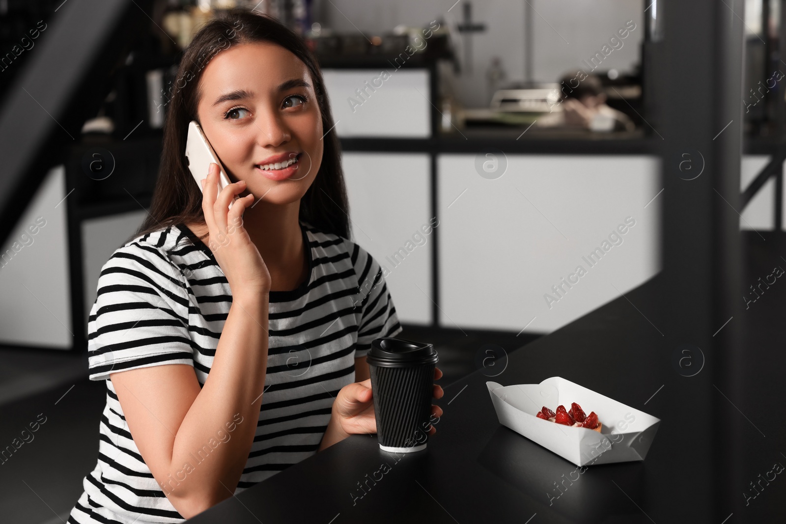 Photo of Happy young woman with paper cup of coffee talking on smartphone at table in hostel dining room