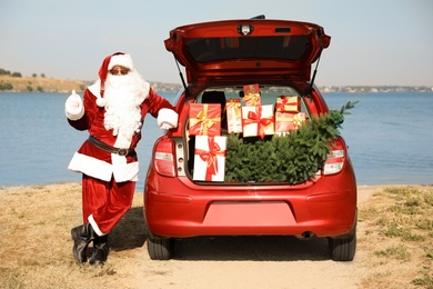 Photo of Authentic Santa Claus near car with open trunk full of presents and fir tree outdoors