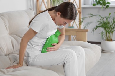 Photo of Young woman using hot water bottle to relieve cystitis pain on sofa at home
