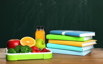 Image of Tray with healthy food and notebooks on wooden table near green chalkboard. School lunch