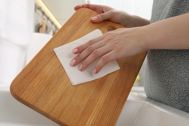 Photo of Woman wiping wooden cutting board with paper napkin at home, closeup