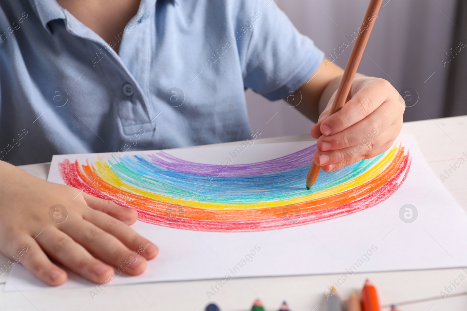 Photo of Little boy drawing rainbow with pencil at white table indoors, closeup. Child`s art