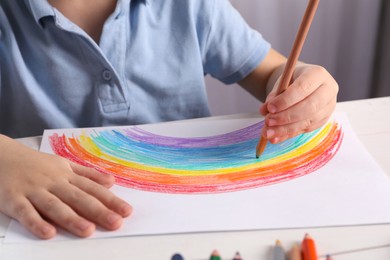 Little boy drawing rainbow with pencil at white table indoors, closeup. Child`s art