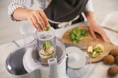 Photo of Young woman putting fresh kiwi into juicer at table in kitchen, closeup