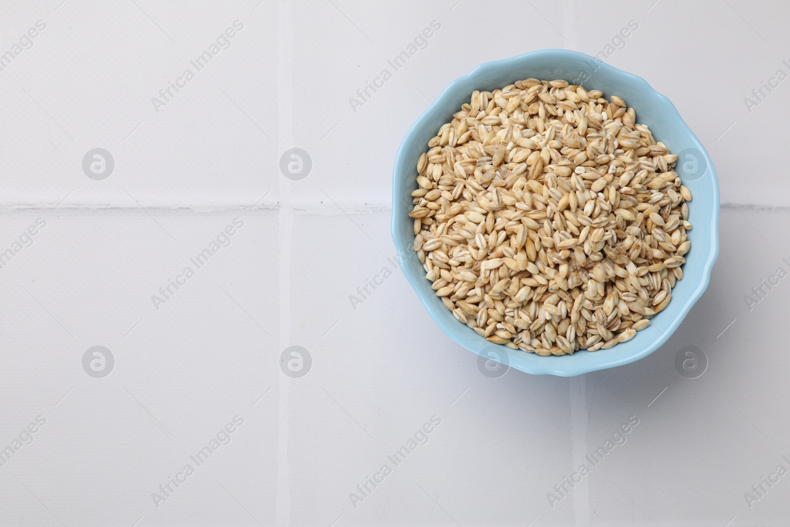Photo of Dry pearl barley in bowl on white tiled table, top view. Space for text