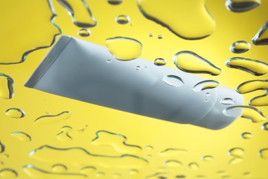 Photo of Moisturizing cream in tube on glass with water drops against yellow background