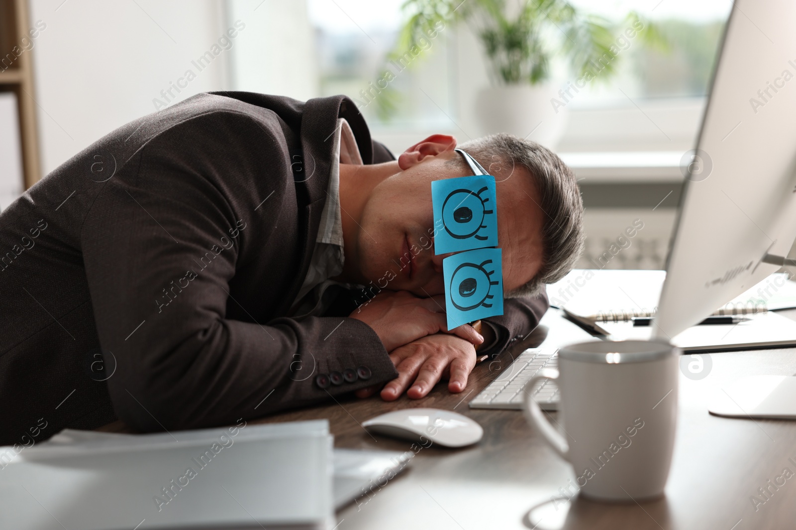 Photo of Man with fake eyes painted on sticky notes snoozing at workplace in office