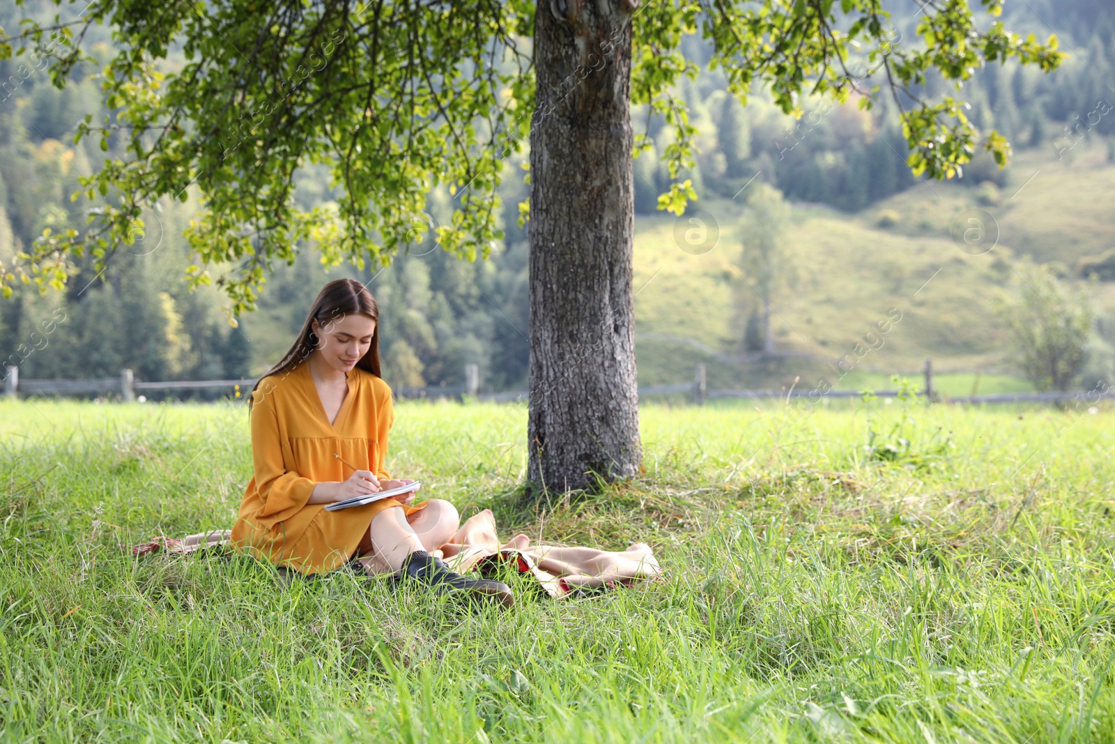 Photo of Beautiful young woman drawing with pencil in notepad on green grass near tree