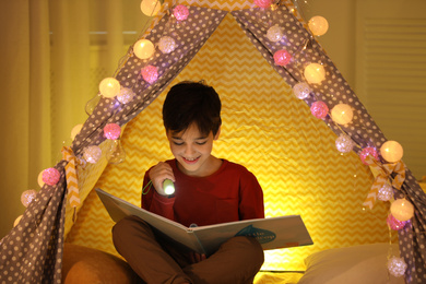 Photo of Boy with flashlight reading book in play tent at home