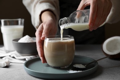 Woman pouring coconut milk into glass of coffee at light grey table, closeup