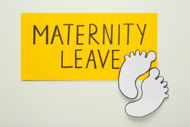 Photo of Note with words Maternity Leave and paper cutout of feet on white background, top view