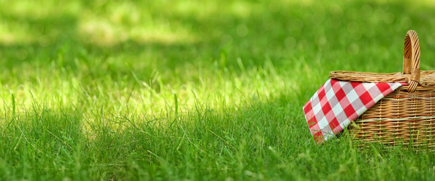 Image of Wicker basket with picnic blanket on green grass in park, space for text. Banner design