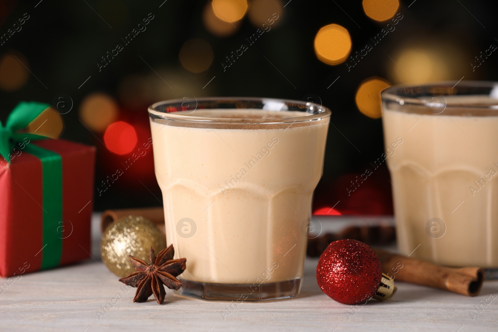 Photo of Tasty eggnog, cinnamon, anise and Christmas decorations on white wooden table against blurred lights