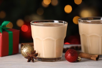 Tasty eggnog, cinnamon, anise and Christmas decorations on white wooden table against blurred lights