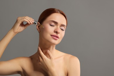 Beautiful young woman applying cosmetic serum onto her face on grey background, space for text