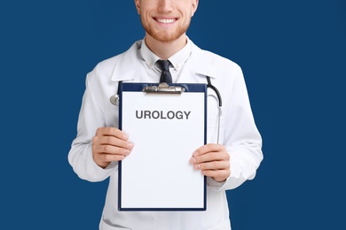 Male doctor holding clipboard with word UROLOGY on blue background, closeup