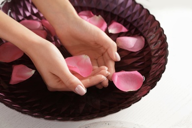 Photo of Woman soaking her hands in bowl with water and petals on table, closeup. Spa treatment