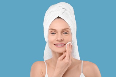 Photo of Beautiful woman in terry towel removing makeup with cotton pad on light blue background