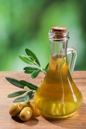 Photo of Jug of cooking oil, olives and green leaves on wooden table against blurred background