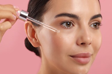 Photo of Beautiful young woman applying serum onto her face on pink background, closeup