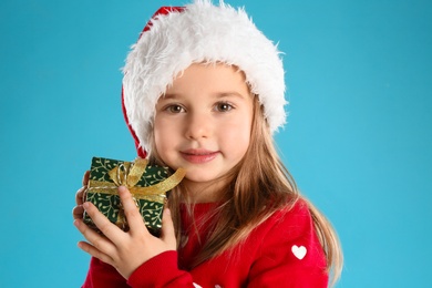 Cute child in Santa hat with Christmas gift on light blue background