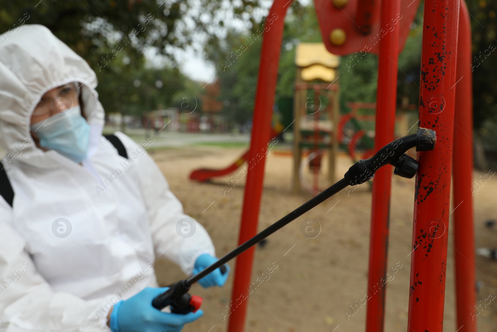 Photo of Woman wearing chemical protective suit at playground, focus on disinfectant sprayer. Preventive measure during coronavirus pandemic