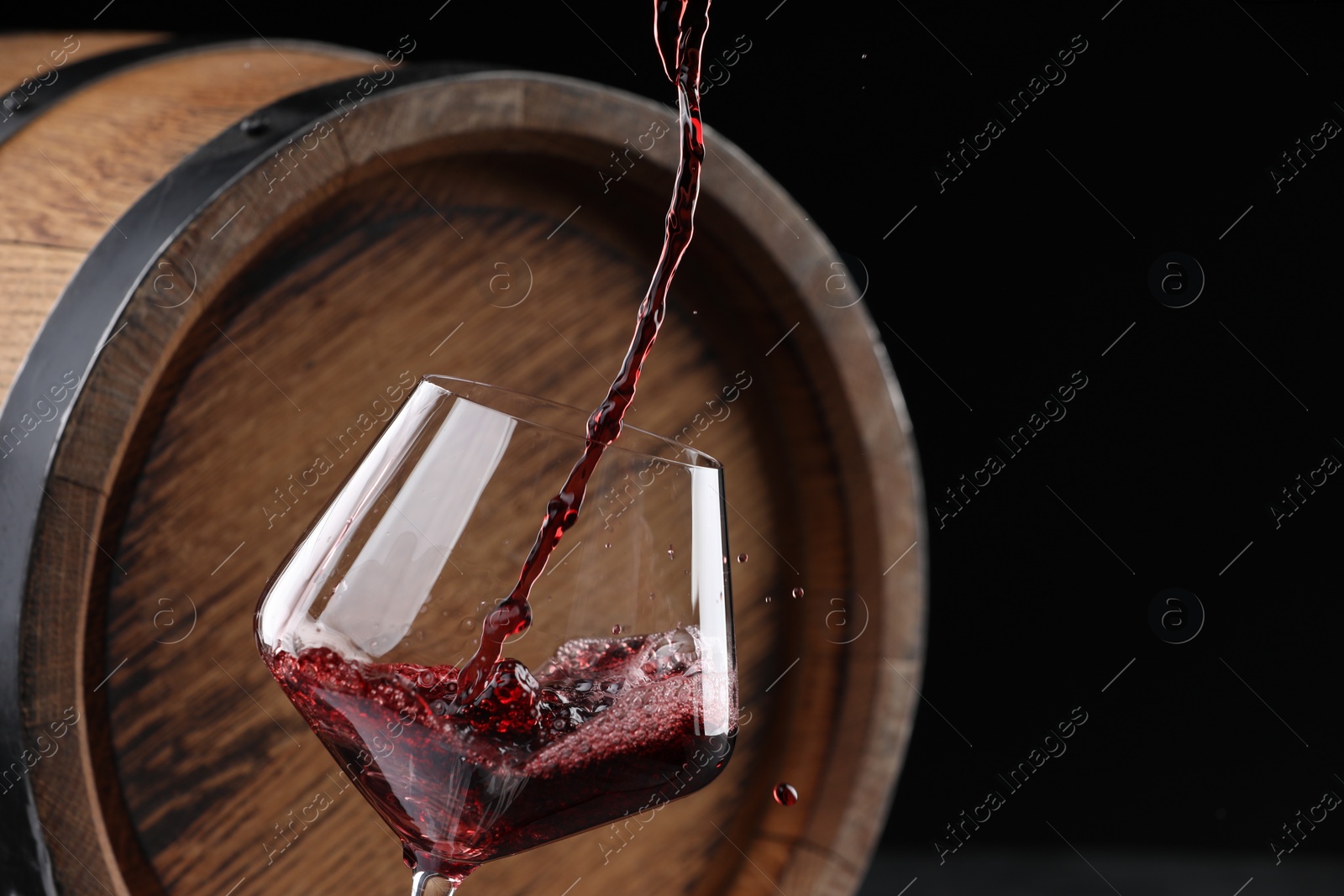 Photo of Pouring delicious wine into glass near wooden barrel against black background, closeup. Space for text